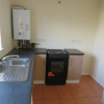 Property for sale by Auction A small kitchen with a black stove and oven in the center, flanked by wood cabinets. A stainless steel sink with a tap is on the left. The kitchen has beige walls, a water heater mounted above, and terracotta tiled flooring. Presented by Palace Auctions