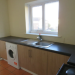 Property for sale by Auction A kitchen with a sink and faucet, a washing machine under the counter, and wooden cabinets. A window above the counter lets in natural light. A glimpse of a living area with a sofa and rug is visible through an arched opening. Presented by Palace Auctions