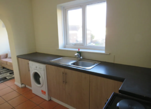 Property for sale by Auction A kitchen with a sink and faucet, a washing machine under the counter, and wooden cabinets. A window above the counter lets in natural light. A glimpse of a living area with a sofa and rug is visible through an arched opening. Presented by Palace Auctions