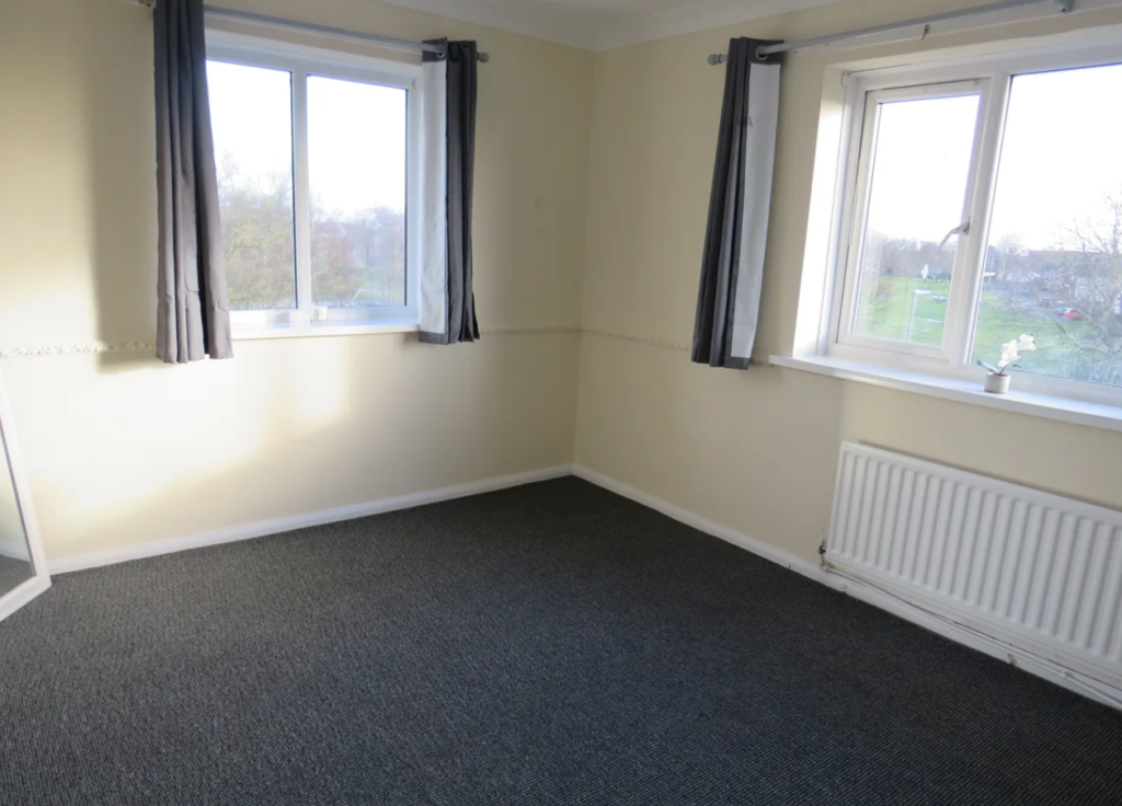 Property for sale by Auction A small empty room with light-colored walls and two windows. The floor is carpeted in dark gray, and there's a white radiator under one window. The windows have dark curtains and offer a view of trees and grass outside. Presented by Palace Auctions