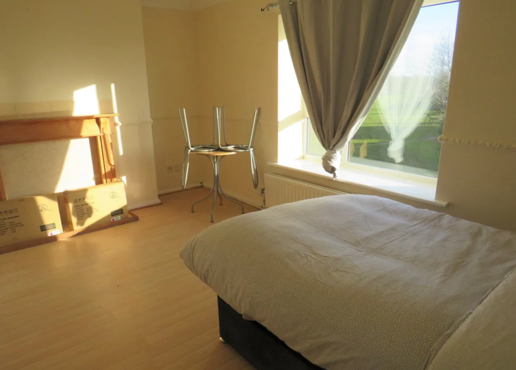 Property for sale by Auction A sunlit bedroom with a neatly made bed, light wood flooring, and a window with curtains drawn to one side. A small round table with chairs upside down on it is near the window. A fireplace with empty boxes is visible on the left. Presented by Palace Auctions