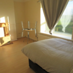 Property for sale by Auction A sunlit bedroom with a neatly made bed, light wood flooring, and a window with curtains drawn to one side. A small round table with chairs upside down on it is near the window. A fireplace with empty boxes is visible on the left. Presented by Palace Auctions