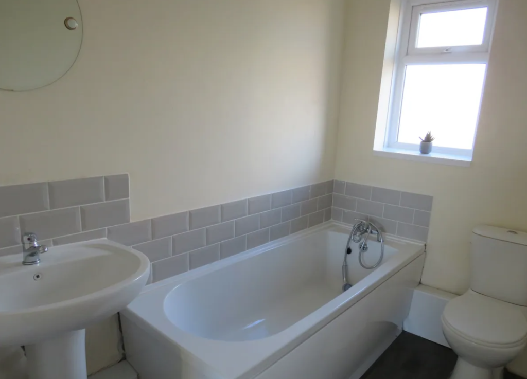Property for sale by Auction A bathroom with a white bathtub, a small basin sink with a round mirror above, and a toilet. The walls have beige tiles around the tub area. A small window with a potted plant sits above the tub. Presented by Palace Auctions