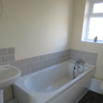 Property for sale by Auction A bathroom with a white bathtub, a small basin sink with a round mirror above, and a toilet. The walls have beige tiles around the tub area. A small window with a potted plant sits above the tub. Presented by Palace Auctions