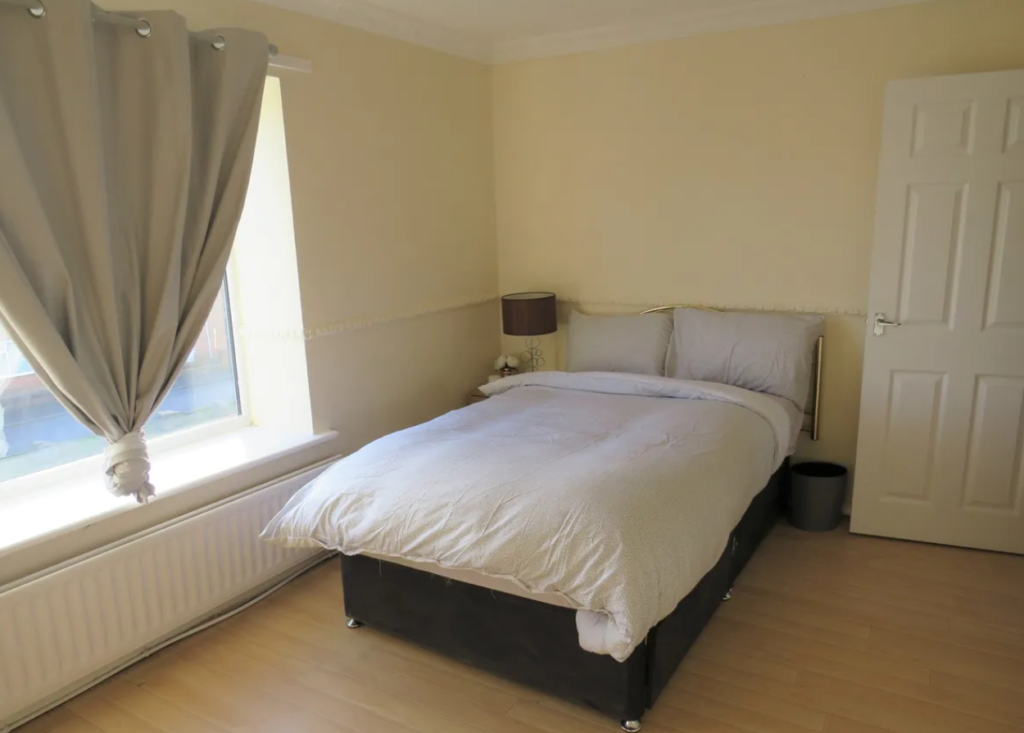 Property for sale by Auction A cozy bedroom with a neatly made bed featuring gray bedding. A small bedside table holds a lamp. The room has beige walls, a large window with gray curtains, and a closed white door. Light wood flooring completes the space. Presented by Palace Auctions