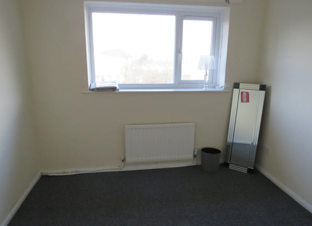 Property for sale by Auction Empty room with beige walls and a gray carpet. A window with a blind is on one wall, with a lamp and some papers on the windowsill. A tall mirror and a wastebasket are positioned against another wall under the window. Presented by Palace Auctions