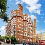 Property for sale by Auction Street view of a large brick building with elegant white details, reminiscent of the luxury apartments for sale in Earls Court, London. Surrounded by trees under a blue sky, a red double-decker bus passes by as parked cars line the road. Presented by Palace Auctions
