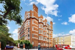 Property for sale by Auction Street view of a large brick building with elegant white details, reminiscent of the luxury apartments for sale in Earls Court, London. Surrounded by trees under a blue sky, a red double-decker bus passes by as parked cars line the road. Presented by Palace Auctions