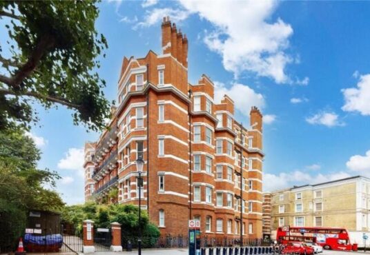 Property for sale by Auction Street view of a large brick building with elegant white details, reminiscent of the luxury apartments for sale in Earls Court, London. Surrounded by trees under a blue sky, a red double-decker bus passes by as parked cars line the road. Presented by Palace Auctions