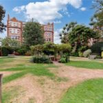 Property for sale by Auction A well-maintained garden with several benches and a central gazebo surrounded by greenery complements the luxury apartments for sale in Earls Court, London. In the background, red brick buildings with pointed roofs rise under a partly cloudy sky on a sunny day. Presented by Palace Auctions