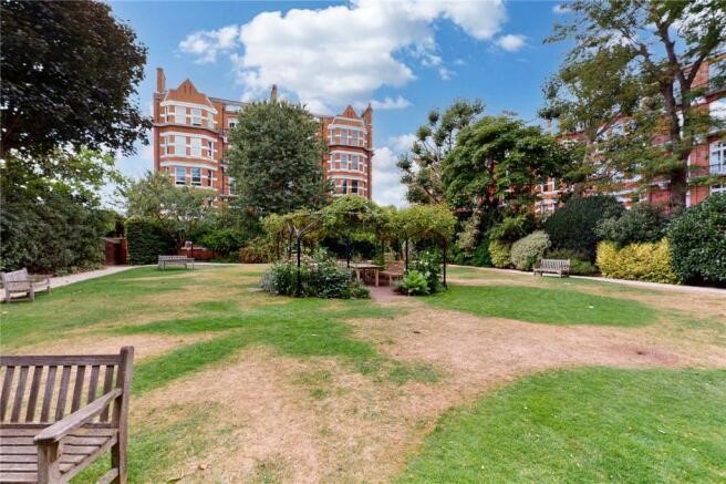 Property for sale by Auction A well-maintained garden with several benches and a central gazebo surrounded by greenery complements the luxury apartments for sale in Earls Court, London. In the background, red brick buildings with pointed roofs rise under a partly cloudy sky on a sunny day. Presented by Palace Auctions