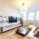 Property for sale by Auction A modern living room in a luxury apartment for sale in Earls Court, London, features a white sofa, wooden coffee table, and large wall-mounted TV. Shelves with decor adorn the walls, while a potted plant graces the bay window beneath a contemporary chandelier. Presented by Palace Auctions