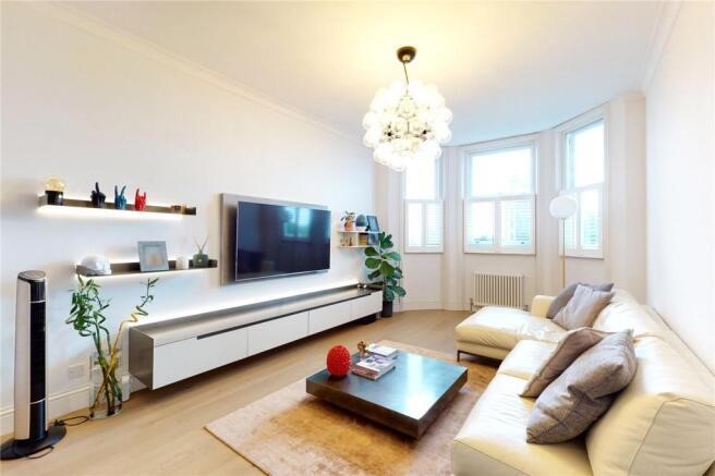 Property for sale by Auction A modern living room in a luxury apartment for sale in Earls Court, London, features a white sofa, wooden coffee table, and large wall-mounted TV. Shelves with decor adorn the walls, while a potted plant graces the bay window beneath a contemporary chandelier. Presented by Palace Auctions