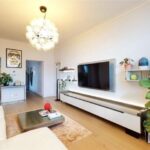 Property for sale by Auction In a luxury apartment for sale in Earls Court, London, the modern living room boasts a white sofa, a large TV on the wall with a sleek console below. Shelves display plants and decor items, while a unique bubble-like ceiling light hangs above. A large green plant graces the corner, complemented by light wood flooring. Presented by Palace Auctions