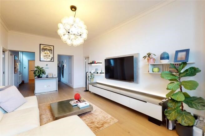 Property for sale by Auction In a luxury apartment for sale in Earls Court, London, the modern living room boasts a white sofa, a large TV on the wall with a sleek console below. Shelves display plants and decor items, while a unique bubble-like ceiling light hangs above. A large green plant graces the corner, complemented by light wood flooring. Presented by Palace Auctions