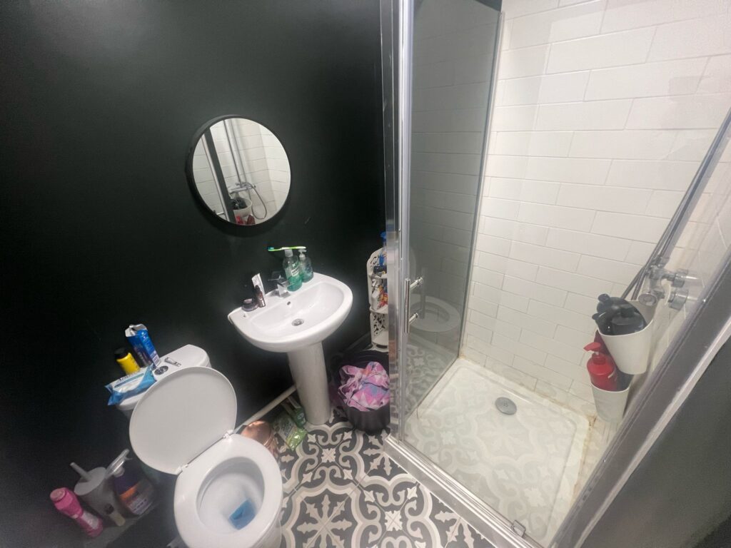 Property for sale by Auction A small bathroom with dark walls features a round mirror above a white sink. There's a shower with a glass door and white tiles. A toilet is beside the sink, and various toiletries are arranged on the sink and toilet tank. The floor has patterned tiles. Presented by Palace Auctions