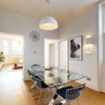 Property for sale by Auction A modern dining room in a luxury apartment for sale in Earls Court, London, features a glass table and four wire chairs on hardwood flooring. A pendant light hangs above the table. The room boasts white walls, artwork, a wall clock, and large windows inviting abundant natural light. Presented by Palace Auctions