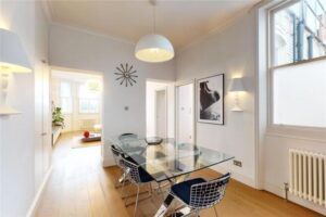 Property for sale by Auction A modern dining room in a luxury apartment for sale in Earls Court, London, features a glass table and four wire chairs on hardwood flooring. A pendant light hangs above the table. The room boasts white walls, artwork, a wall clock, and large windows inviting abundant natural light. Presented by Palace Auctions