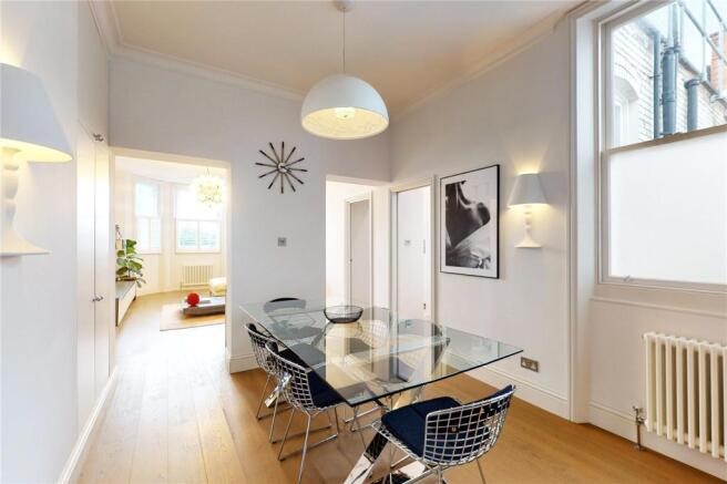 Property for sale by Auction A modern dining room in a luxury apartment for sale in Earls Court, London, features a glass table and four wire chairs on hardwood flooring. A pendant light hangs above the table. The room boasts white walls, artwork, a wall clock, and large windows inviting abundant natural light. Presented by Palace Auctions