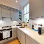 Property for sale by Auction In a luxury apartment for sale in Earl's Court, London, discover a compact kitchen with gray cabinets and a white countertop. It features a gas stove with an oven, a sink, and small appliances like a toaster and coffee maker. A towel hangs on the oven door as natural light floods through the window. Presented by Palace Auctions