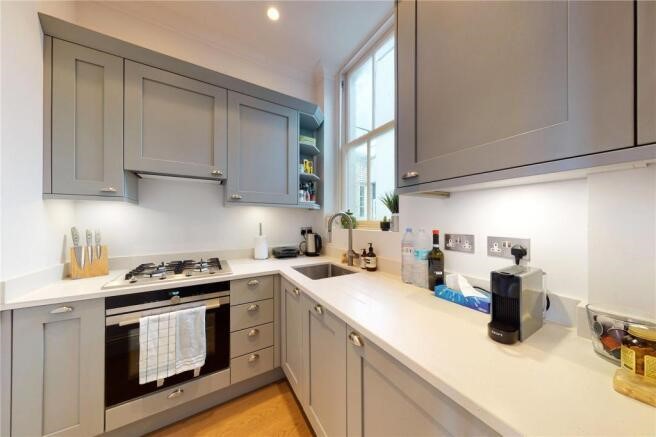 Property for sale by Auction In a luxury apartment for sale in Earl's Court, London, discover a compact kitchen with gray cabinets and a white countertop. It features a gas stove with an oven, a sink, and small appliances like a toaster and coffee maker. A towel hangs on the oven door as natural light floods through the window. Presented by Palace Auctions