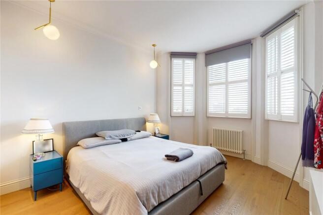 Property for sale by Auction This luxury apartment for sale in Earls Court, London features a bright bedroom with a large bed, two blue nightstands with lamps, and wall-mounted globe lights. Three tall windows with shutters flood the space with natural light. A radiator and garment rack complete the elegant setting. Presented by Palace Auctions