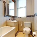 Property for sale by Auction A luxury apartment for sale in Earls Court, London, features a small bathroom with a bathtub on the left, a sink with a cabinet and mirror above, and a toilet on the right. The walls boast patterned tiles, complemented by a frosted window above the sink and toilet. A towel hangs neatly on a ring holder. Presented by Palace Auctions