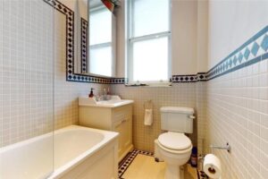 Property for sale by Auction A luxury apartment for sale in Earls Court, London, features a small bathroom with a bathtub on the left, a sink with a cabinet and mirror above, and a toilet on the right. The walls boast patterned tiles, complemented by a frosted window above the sink and toilet. A towel hangs neatly on a ring holder. Presented by Palace Auctions
