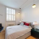 Property for sale by Auction A bright bedroom in a luxury apartment for sale in Earls Court, London, features a queen-sized bed with gray bedding and red pillows. Two bedside tables hold lamps with white shades. Three pendant lights hang from the ceiling, and a window with white shutters lets in natural light. A chair sits in the corner. Presented by Palace Auctions