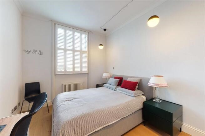 Property for sale by Auction A bright bedroom in a luxury apartment for sale in Earls Court, London, features a queen-sized bed with gray bedding and red pillows. Two bedside tables hold lamps with white shades. Three pendant lights hang from the ceiling, and a window with white shutters lets in natural light. A chair sits in the corner. Presented by Palace Auctions