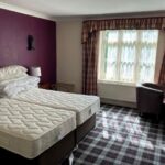 Property for sale by Auction A hotel room with two twin beds pushed together, topped with multiple pillows. The room has a plaid carpet, plum-colored accent wall, and a window with checkered curtains. There are two armchairs and a round table near the window. Presented by Palace Auctions