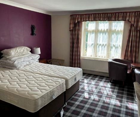 Property for sale by Auction A hotel room with two twin beds pushed together, topped with multiple pillows. The room has a plaid carpet, plum-colored accent wall, and a window with checkered curtains. There are two armchairs and a round table near the window. Presented by Palace Auctions