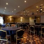 Property for sale by Auction A dimly lit conference room with multiple round tables covered in dark cloths, each surrounded by chairs. The room features decorative lighting fixtures, patterned carpet, and framed artwork on the walls. A large screen is visible at the front. Presented by Palace Auctions