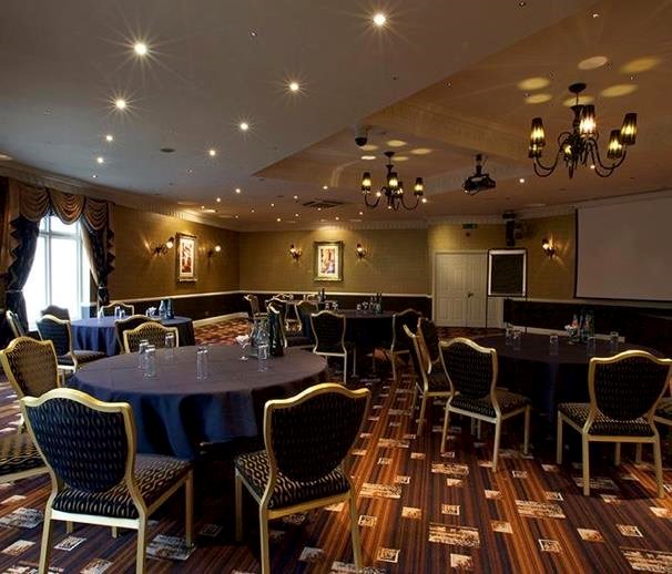 Property for sale by Auction A dimly lit conference room with multiple round tables covered in dark cloths, each surrounded by chairs. The room features decorative lighting fixtures, patterned carpet, and framed artwork on the walls. A large screen is visible at the front. Presented by Palace Auctions