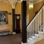 Property for sale by Auction An elegant hallway with patterned carpet, a white staircase, and a tall grandfather clock. A portrait hangs on the wall above an ornate bench with gold detailing. Arched ceilings and two hanging lanterns are visible. Presented by Palace Auctions