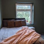 Property for sale by Auction A neatly made bed with brown-patterned pillows and a pink blanket is in a small room with green walls. A window, partially covered by the curtain, reveals a view of trees outside. A few small items are on the window sill. Presented by Palace Auctions