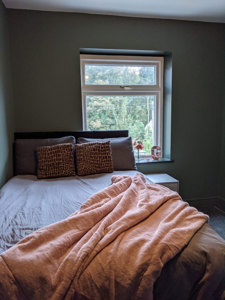Property for sale by Auction A neatly made bed with brown-patterned pillows and a pink blanket is in a small room with green walls. A window, partially covered by the curtain, reveals a view of trees outside. A few small items are on the window sill. Presented by Palace Auctions