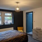 Property for sale by Auction Bedroom with a modern design featuring a bed with geometric patterned bedding, a yellow chair by the window, and a gray dresser with a decorative box. The room has gray walls, a hanging light fixture, and a doorway leading to another room. Presented by Palace Auctions