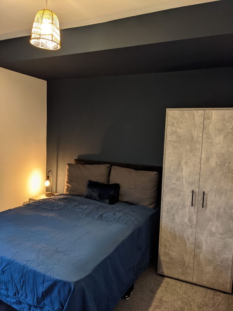 Property for sale by Auction A modern bedroom with a dark blue accent wall, a bed dressed in a blue cover, and gray pillows. A small bedside table with a lamp is on the left. A textured gray wardrobe is on the right. A rattan pendant light hangs from the ceiling. Presented by Palace Auctions