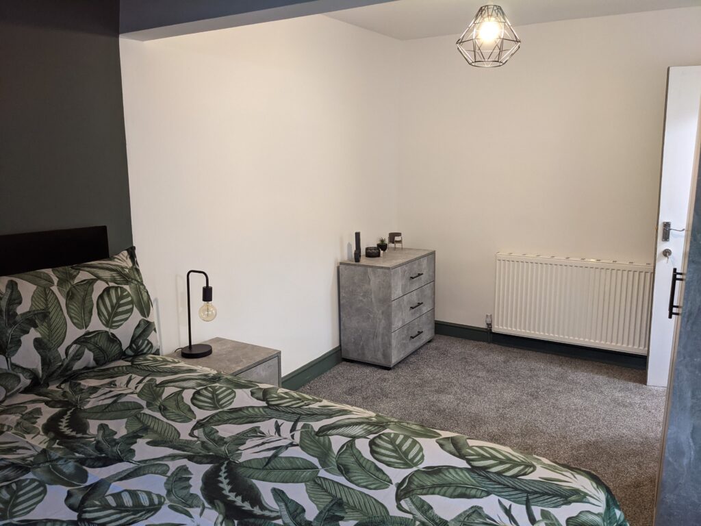 Property for sale by Auction A modern bedroom with tropical leaf-patterned bedding on a bed. A geometric pendant light hangs from the ceiling. A gray dresser and a bedside table with a lamp are placed against the walls. The room has gray carpet flooring and a radiator. Presented by Palace Auctions