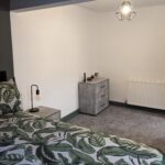 Property for sale by Auction A modern bedroom with tropical leaf-patterned bedding on a bed. A geometric pendant light hangs from the ceiling. A gray dresser and a bedside table with a lamp are placed against the walls. The room has gray carpet flooring and a radiator. Presented by Palace Auctions