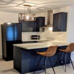 Property for sale by Auction Modern kitchen with black cabinets and appliances. A marble island counter features two brown barstools. Overhead is a stylish geometric light fixture. White subway tiles line the backsplash, and the floor is tiled in gray. Presented by Palace Auctions