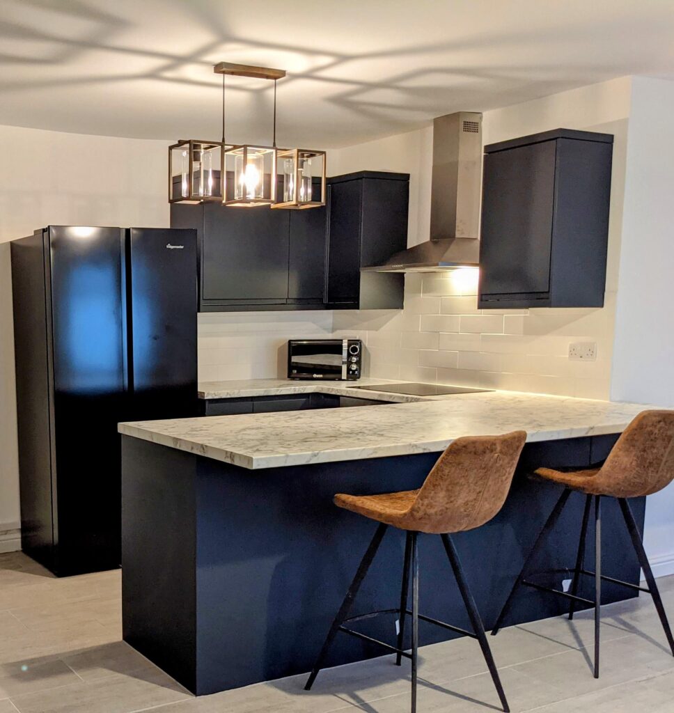 Property for sale by Auction Modern kitchen with black cabinets and appliances. A marble island counter features two brown barstools. Overhead is a stylish geometric light fixture. White subway tiles line the backsplash, and the floor is tiled in gray. Presented by Palace Auctions