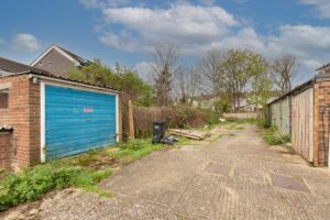 Property for sale by Auction A narrow alley with old brick garages on both sides, featuring weathered blue and green doors. Overgrown weeds and scattered debris are visible around, with a partly cloudy sky overhead. Presented by Palace Auctions