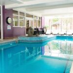 Property for sale by Auction Indoor swimming pool with turquoise tiles, surrounded by purple walls. Lounge chairs line the windows, and a clock hangs on the wall. Large windows provide a view of an outdoor area with greenery and brick buildings. Presented by Palace Auctions