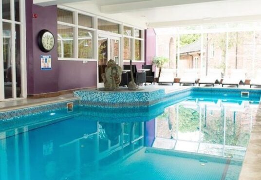 Property for sale by Auction Indoor swimming pool with turquoise tiles, surrounded by purple walls. Lounge chairs line the windows, and a clock hangs on the wall. Large windows provide a view of an outdoor area with greenery and brick buildings. Presented by Palace Auctions