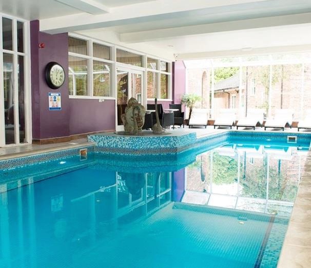 Property for sale by Auction Indoor swimming pool with turquoise tiles, surrounded by purple walls. Lounge chairs line the windows, and a clock hangs on the wall. Large windows provide a view of an outdoor area with greenery and brick buildings. Presented by Palace Auctions