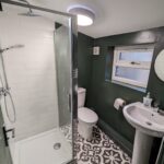 Property for sale by Auction A compact bathroom featuring a glass shower with a rain showerhead, a toilet, a pedestal sink with soap and a circular mirror above. The walls are dark green, and the floor has a black-and-white patterned tile design. Presented by Palace Auctions