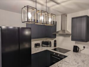 Property for sale by Auction Modern kitchen with black appliances, including a fridge, stove, and microwave. Marble countertops and white tile backsplash enhance the sleek design. Three hanging pendant lights illuminate the space, and a black kettle and knife set are visible. Presented by Palace Auctions