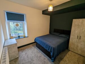 Property for sale by Auction A bedroom with a double bed covered in dark blue bedding. There's a window with a blue blind, a small table beneath it with decor, a pendant light above, and a wardrobe beside the bed. The walls are a mix of white and dark colors, and the carpet is gray. Presented by Palace Auctions